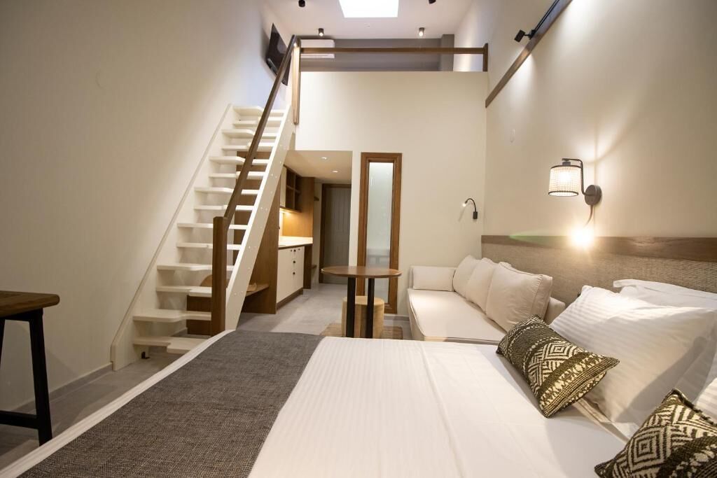 Quarto com cama, almofadas, mesa de cabeceira, mesa, sofá, escada e luminária na parede.