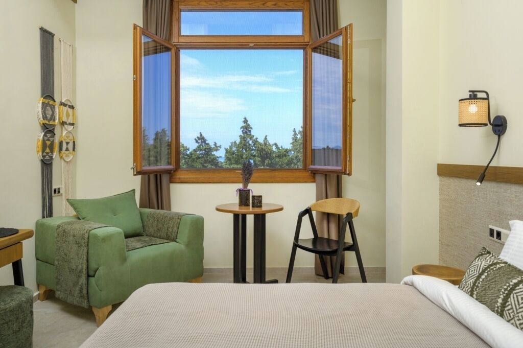 Una habitación con una cama, un sillón verde, una mesita redonda, un ventanal de madera y una lámpara de pared.