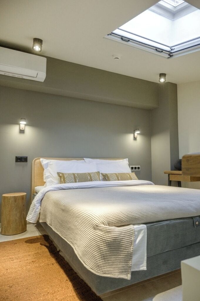 Une chambre à coucher moderne avec un puits de lumière, un lit avec un matelas gris, du linge de lit blanc et une table de chevet en bois.
