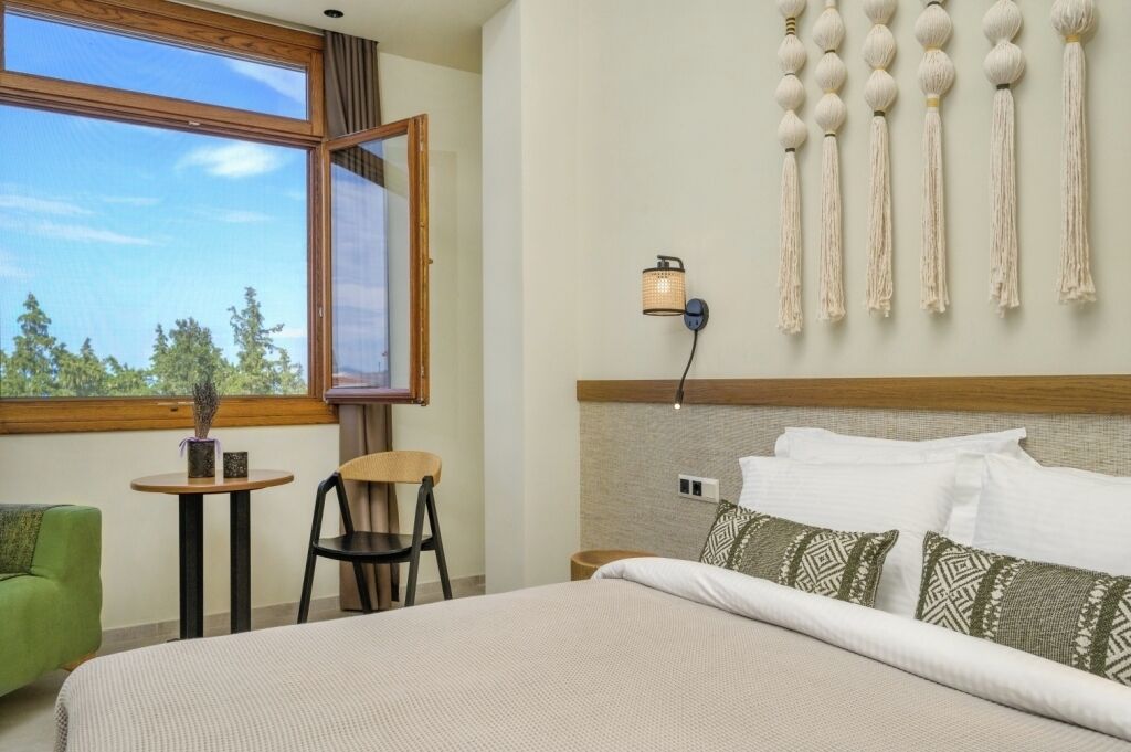 Habitación de hotel con cama, una mesita, una silla, lámpara de pared y ventana con vista a árboles.