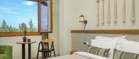 Habitación de hotel con cama, una mesita, una silla, lámpara de pared y ventana con vista a árboles.