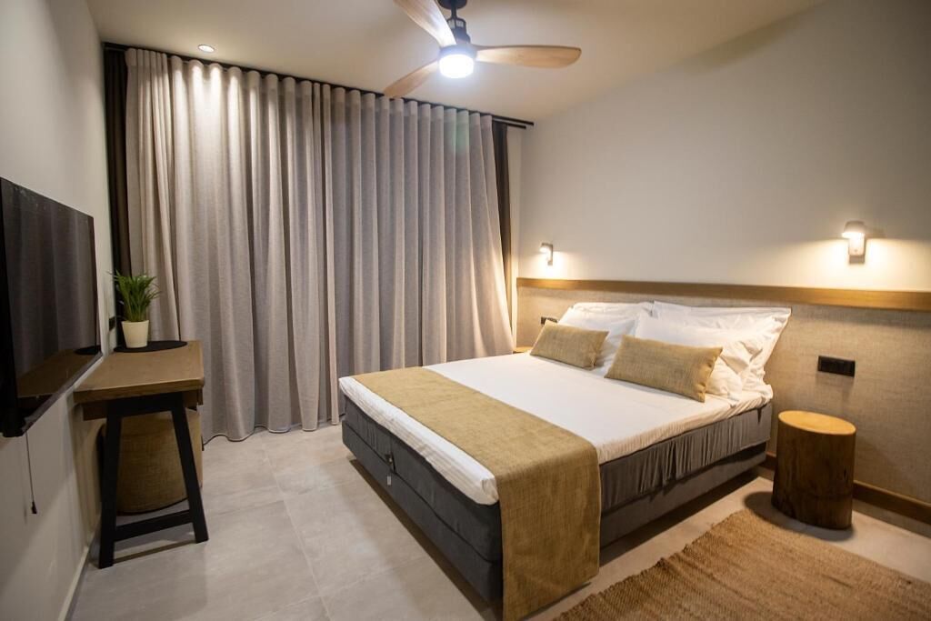 Une chambre à coucher comprenant un lit, un bureau, une chaise, une plante en pot, un ventilateur de plafond et un tabouret rond en bois.