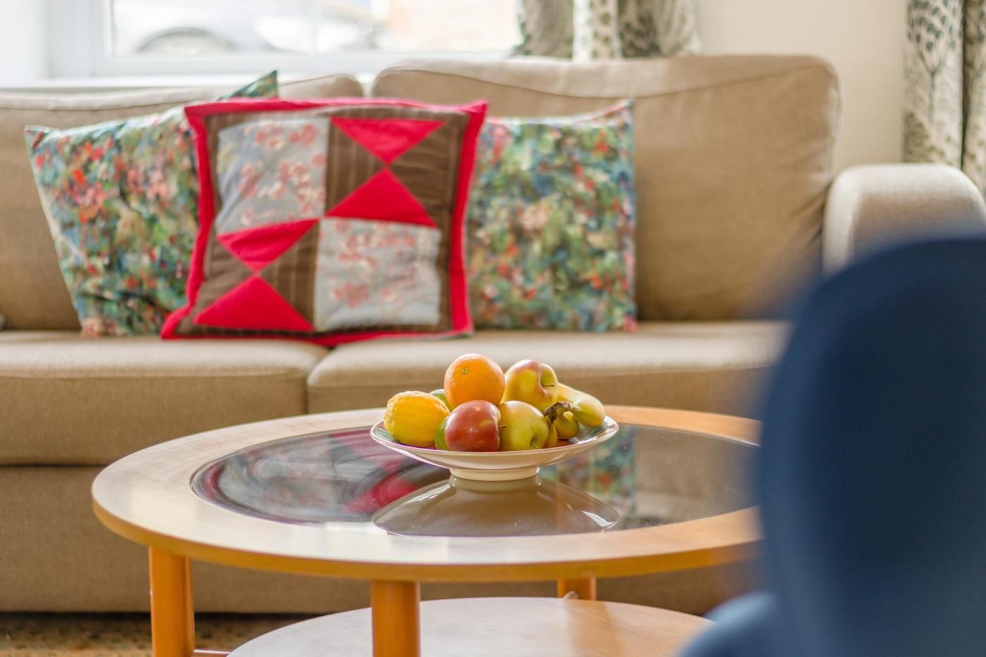 Un salon avec un canapé, une table basse en verre sur laquelle se trouvent des fruits, et un coussin rouge et marron.