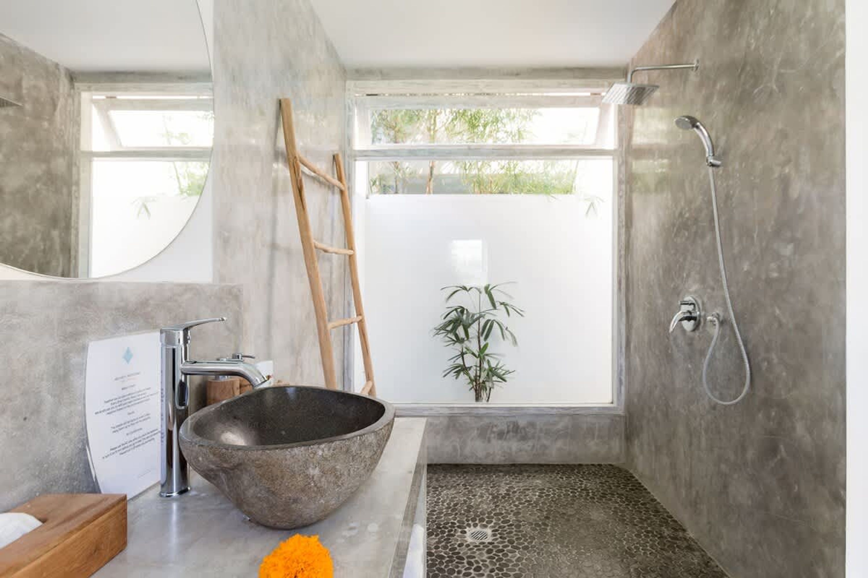 A modern bathroom with a large mirror, a stone basin, a shower, and a view of greenery outside.