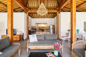 A spacious living area with a wooden ceiling, a sofa, a coffee table with flowers, and a kitchen in the background. - Exquisite Architectural Villa with Outdoor Pool (Seminyak)