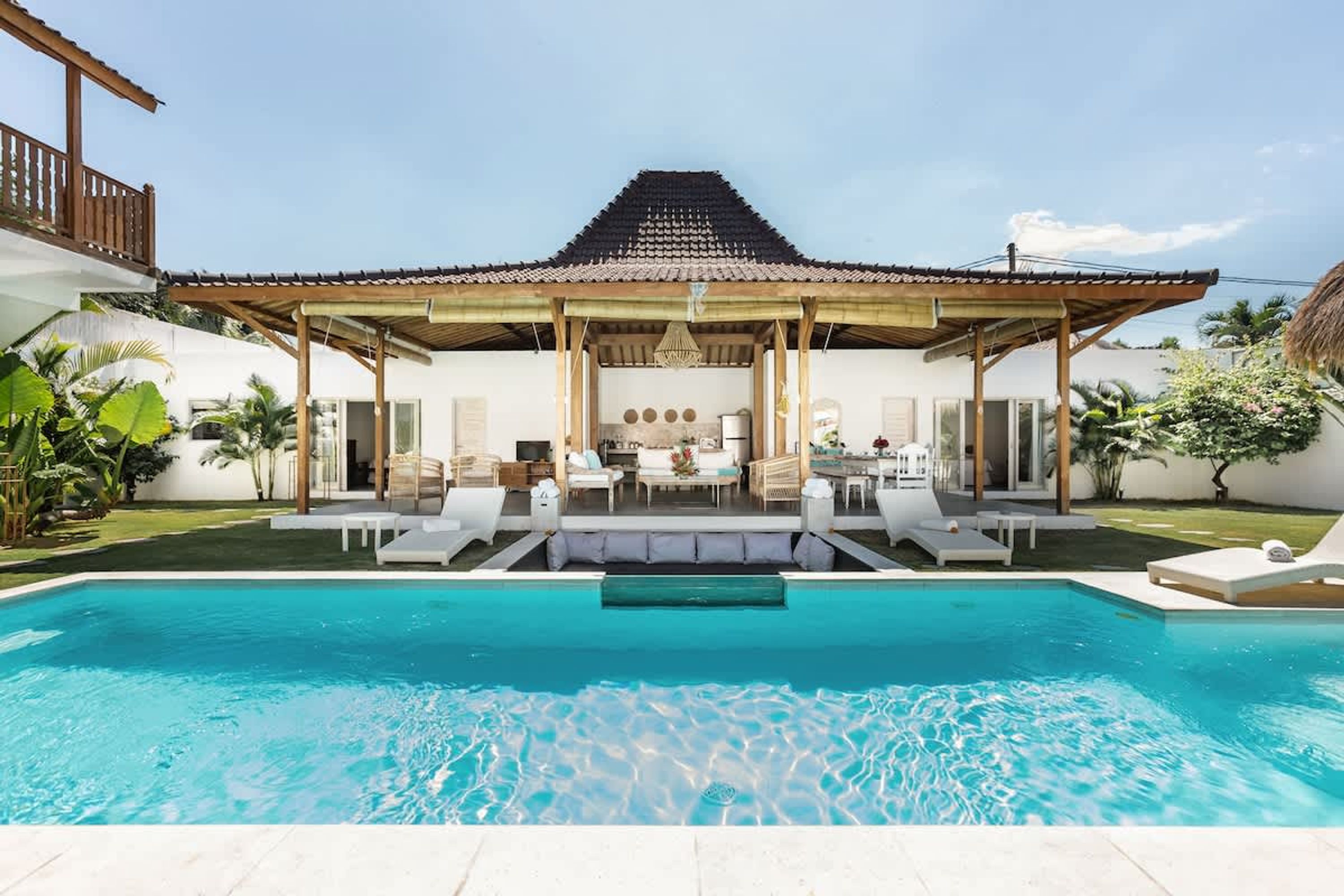 A poolside view of a house with a covered patio, a dining area, and a kitchen visible through the open space.