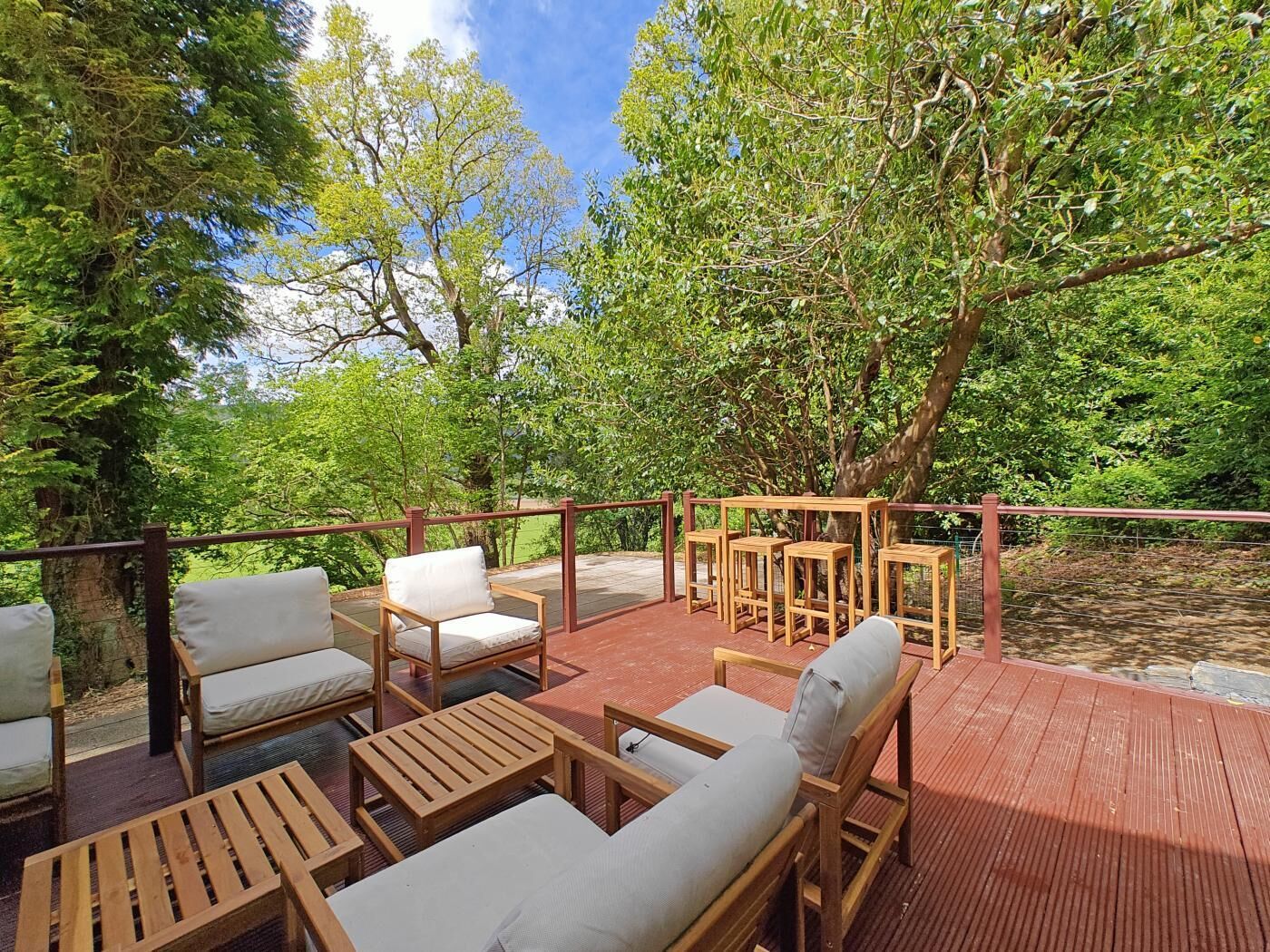 Une terrasse extérieure aménagée avec du mobilier en bois, notamment des chaises et des tables, entourée d’une végétation luxuriante et d’arbres.