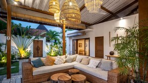 Un espacio al aire libre acogedor con un sofá de mimbre, taburetes de madera y una planta grande en maceta.