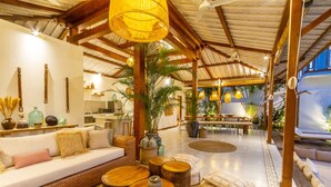 Amplio espacio interior con techo alto, vigas de madera, un conjunto de sofás, mesas redondas de madera y una zona de comedor con mesa y sillas.