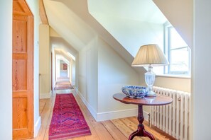 Un couloir avec une porte en bois, un tapis rouge à motifs, une table avec une lampe et un bol, et une fenêtre donnant sur un paysage.