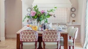 Ein Essbereich mit einem Tisch, der für vier Personen gedeckt ist, umgeben von stuhlförmigen Sitzgelegenheiten mit Blumenmuster. Auf dem Tisch steht eine Vase mit Blumen, sowie Teller und eine Karaffe.