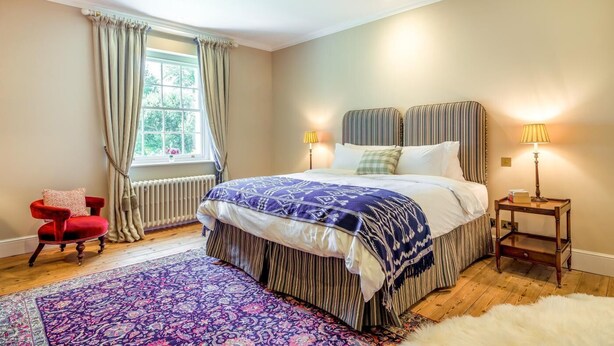 Ein Schlafzimmer mit einem großen Bett, zwei Nachttischlampen, einem Fenster mit Vorhängen, einem roten Sessel und einem gemusterten Teppich.
