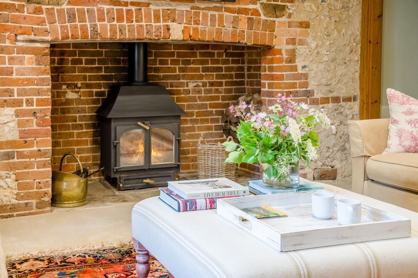A cozy living room with a brick fireplace, a white sofa, a coffee table with books and cups, and a vase with flowers.