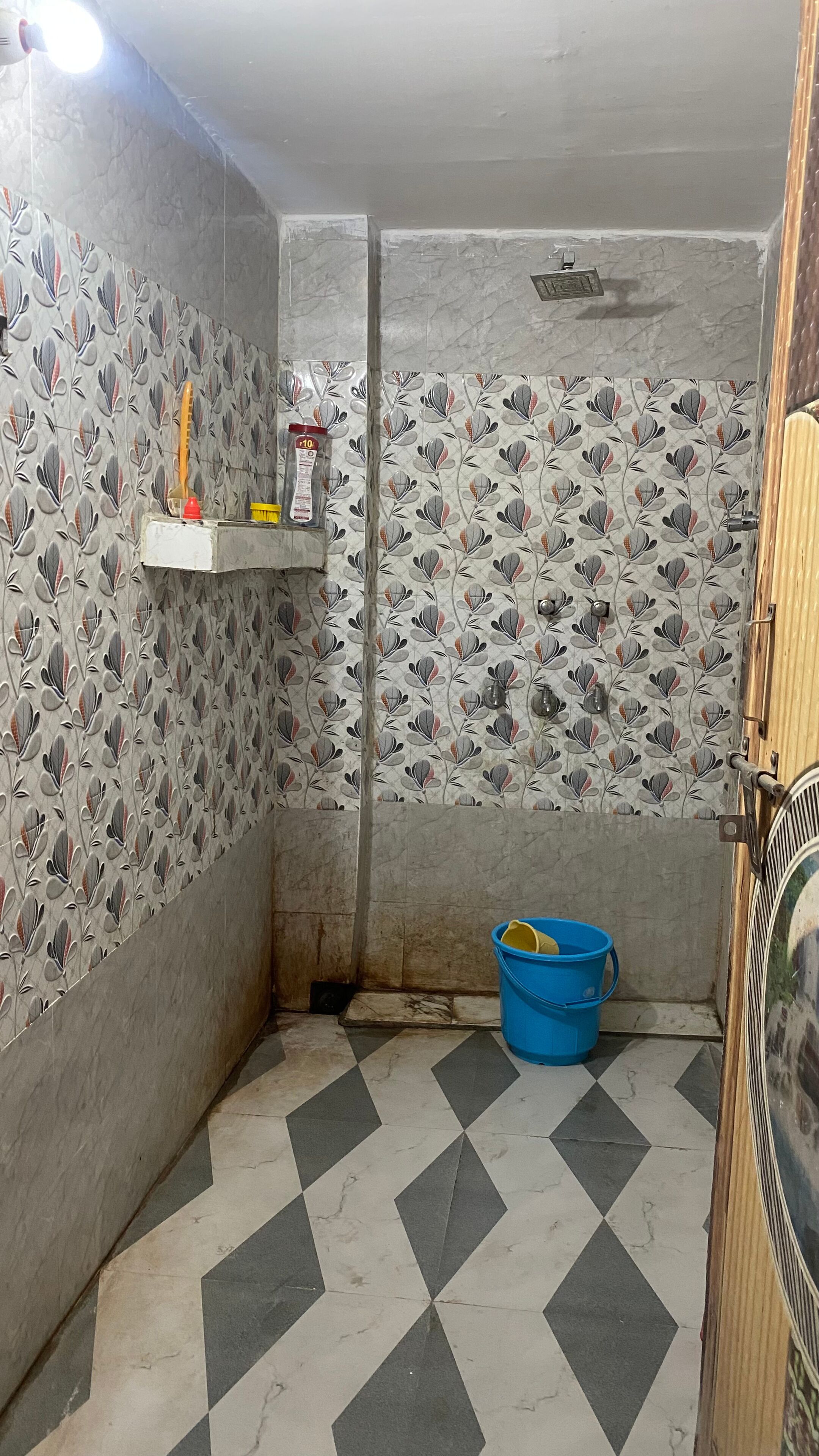 A bathroom with patterned tiles, a blue bucket, and a shelf with cleaning supplies.