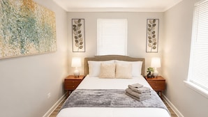 A bedroom with a bed, two bedside tables, a large artwork on the wall, and a window with blinds. - Spacious 5BD 2BA Home Close To Oakbrook (Westmont)