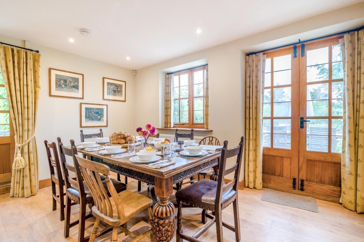 Une salle à manger avec une table en bois dressée pour quatre personnes, entourée de chaises, avec une fenêtre et une porte en arrière-plan.
