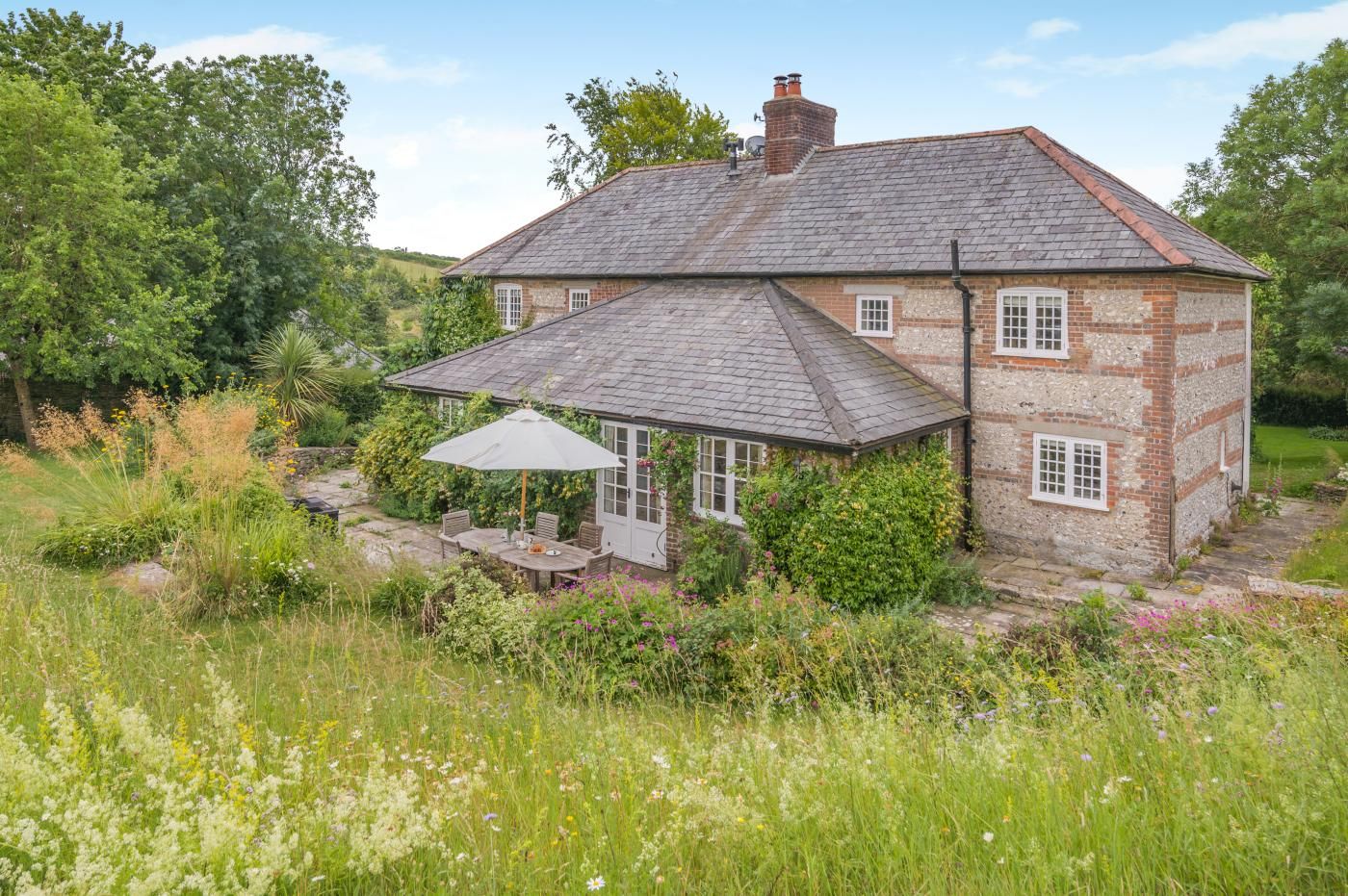 A traditional stone house with a sloped roof, a covered patio with a table and chairs, and a garden with various plants and flowers.
