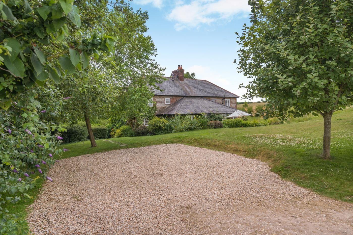 A gravel driveway leading to a stone house with a sloped roof, surrounded by lush greenery and trees.