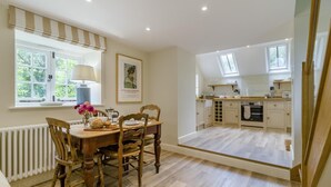 A dining area with a wooden table and chairs, a window with a view of greenery, and a kitchen with cabinets and a countertop. - 1 Bed in Chebbard (oc-p43271) (Puddletown)