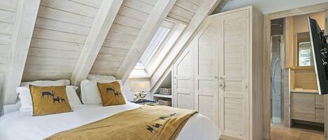 A bedroom with a sloped ceiling, a bed with white linens, a brown throw blanket, and a nightstand with a lamp.