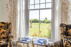 A room with two chairs facing a large window, a small table with a cup and a bowl, and a view of a garden with statues. - 2 Bed in Beaumaris (oc-l44702) (Beaumaris)