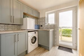 Eine kleine Küche mit grauen Schränken, einer Waschmaschine und einem Fenster mit Blick auf Grünflächen.