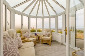 A conservatory with wicker furniture, a small table, and a view of the sea. - 3 Bed in Mundesley (oc-s44224) (Mundesley)