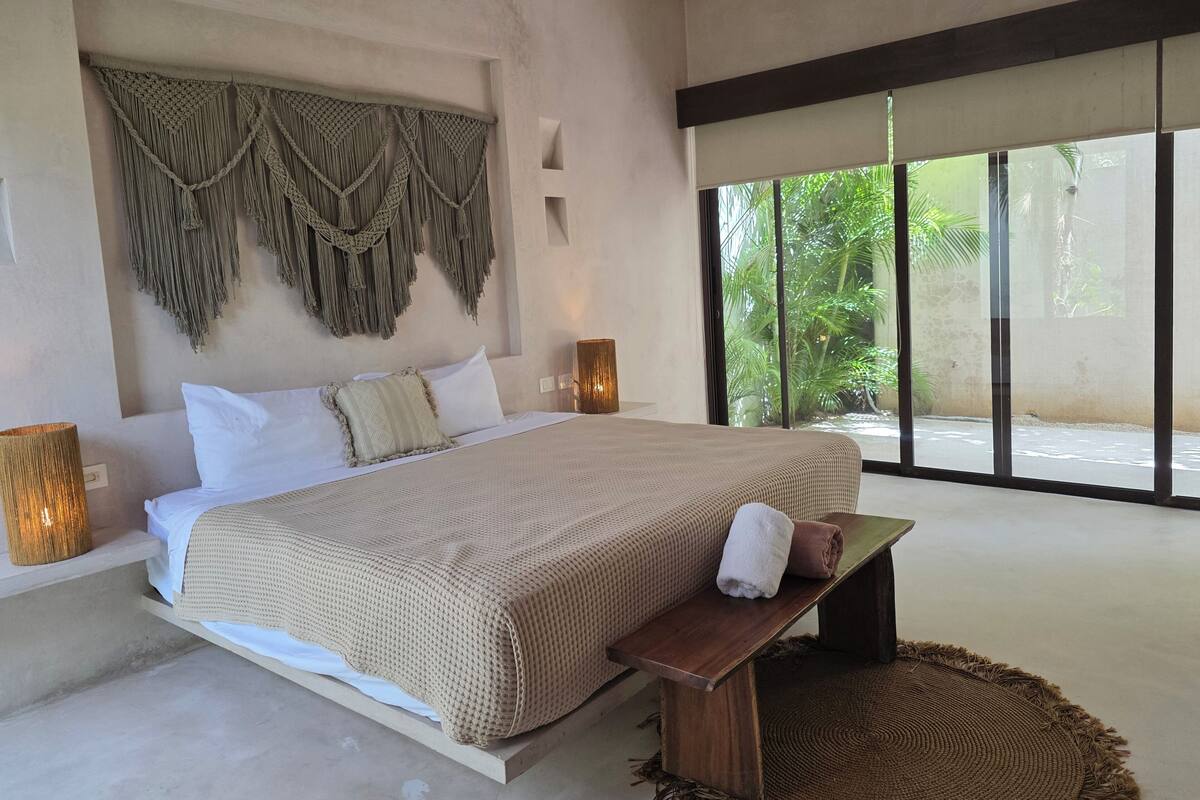 Un dormitorio moderno con una cama grande, un banco y vistas al verdor exterior.