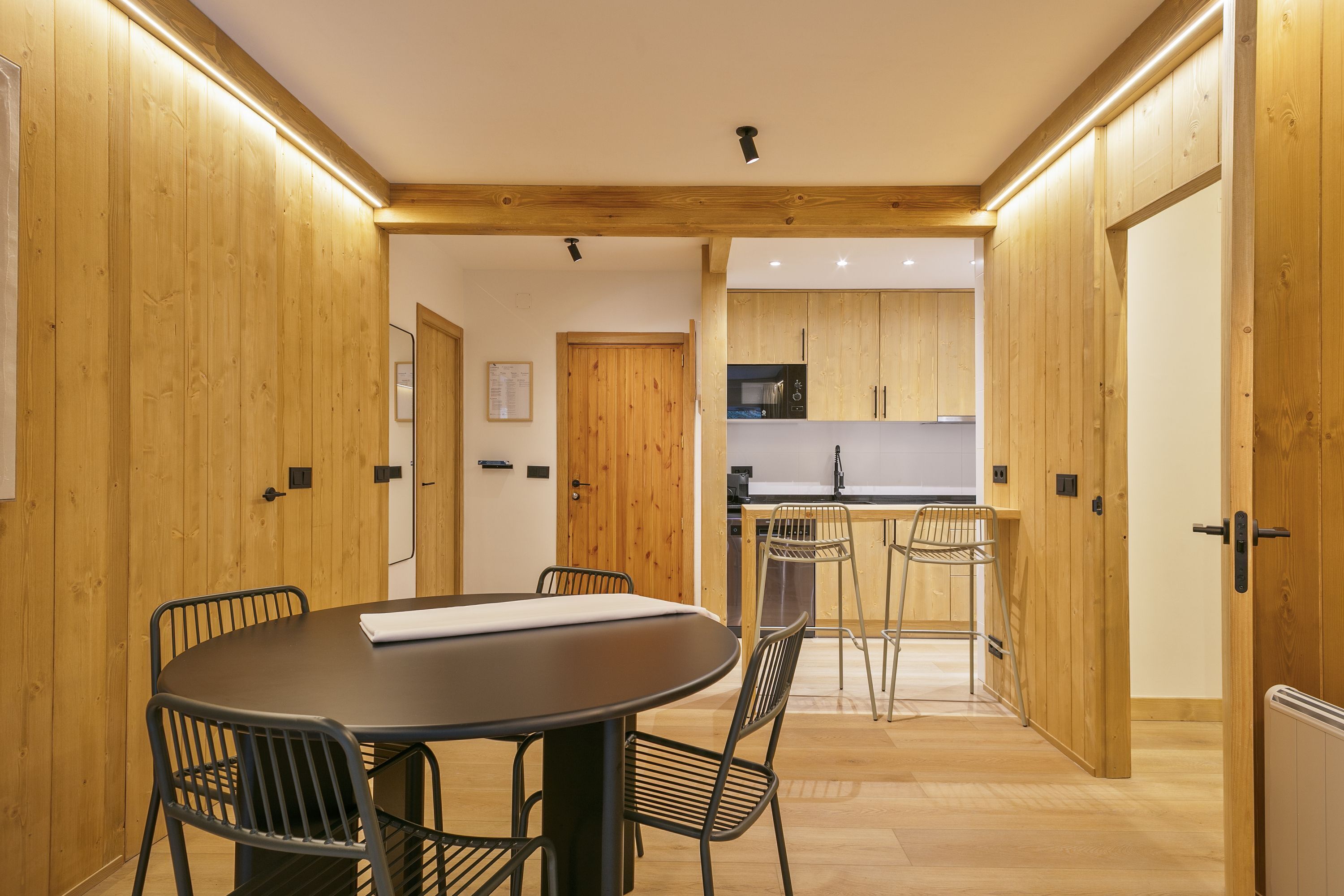 A modern interior with a wooden-paneled wall, a round dining table with chairs, a kitchen area with wooden cabinets, and a visible door.