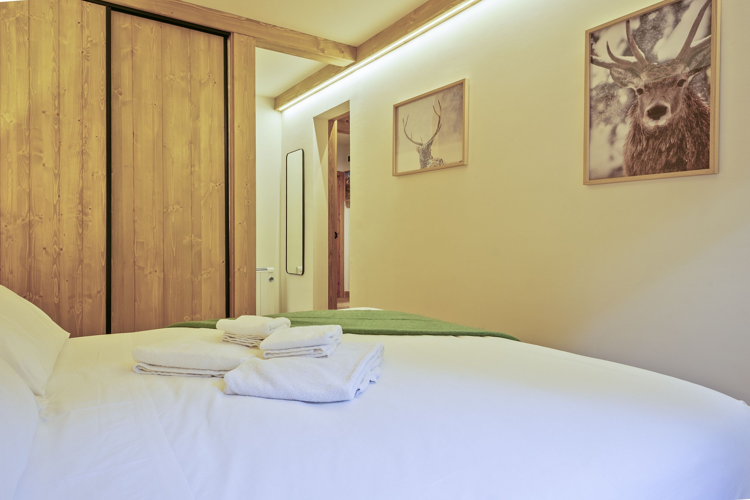 A hotel room with a bed, white linens, a green throw, a wooden wardrobe, and two framed deer head pictures.