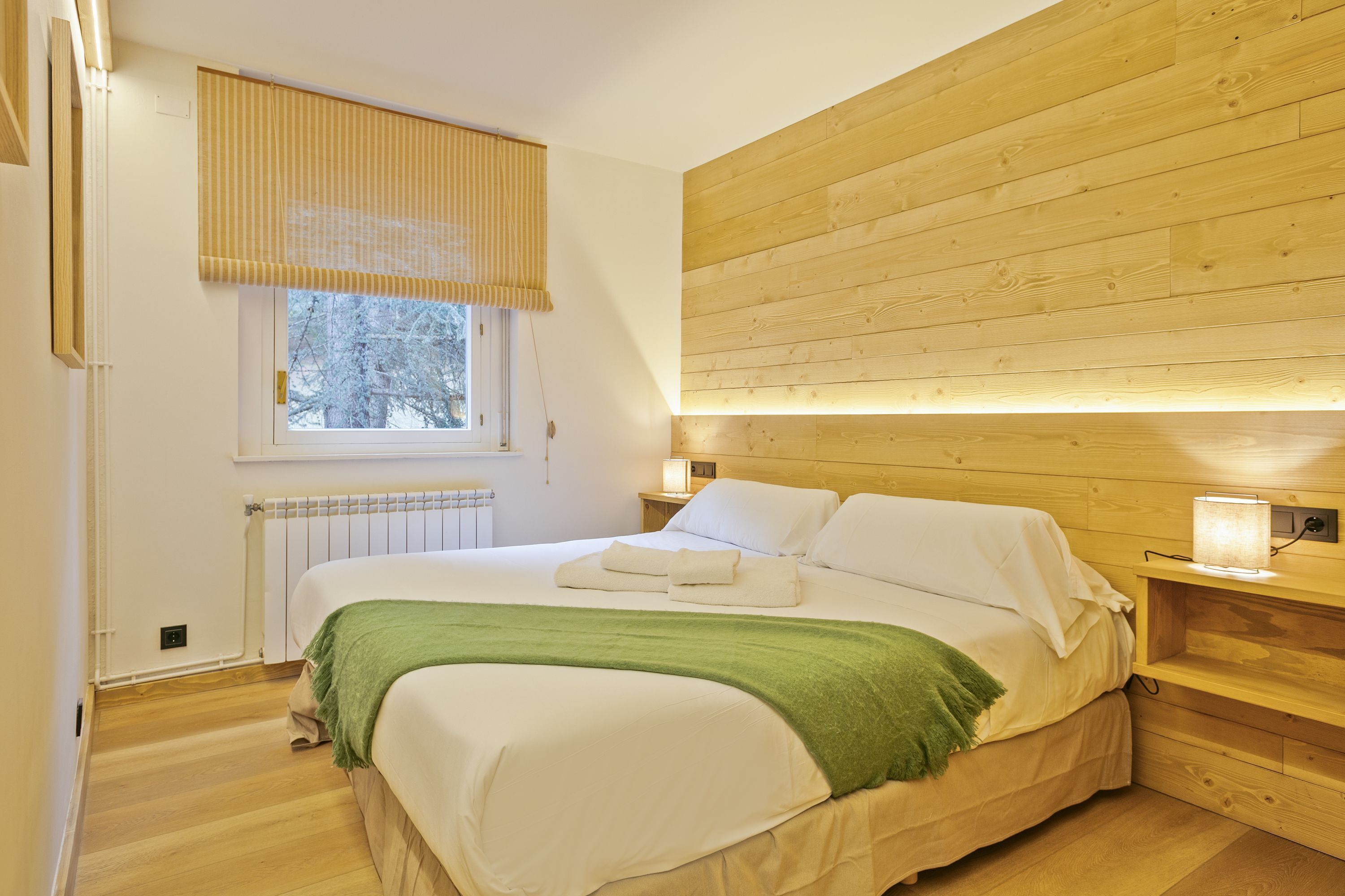 Une chambre à coucher avec une tête de lit en bois, une fenêtre avec des stores, un radiateur et des lampes de chevet.