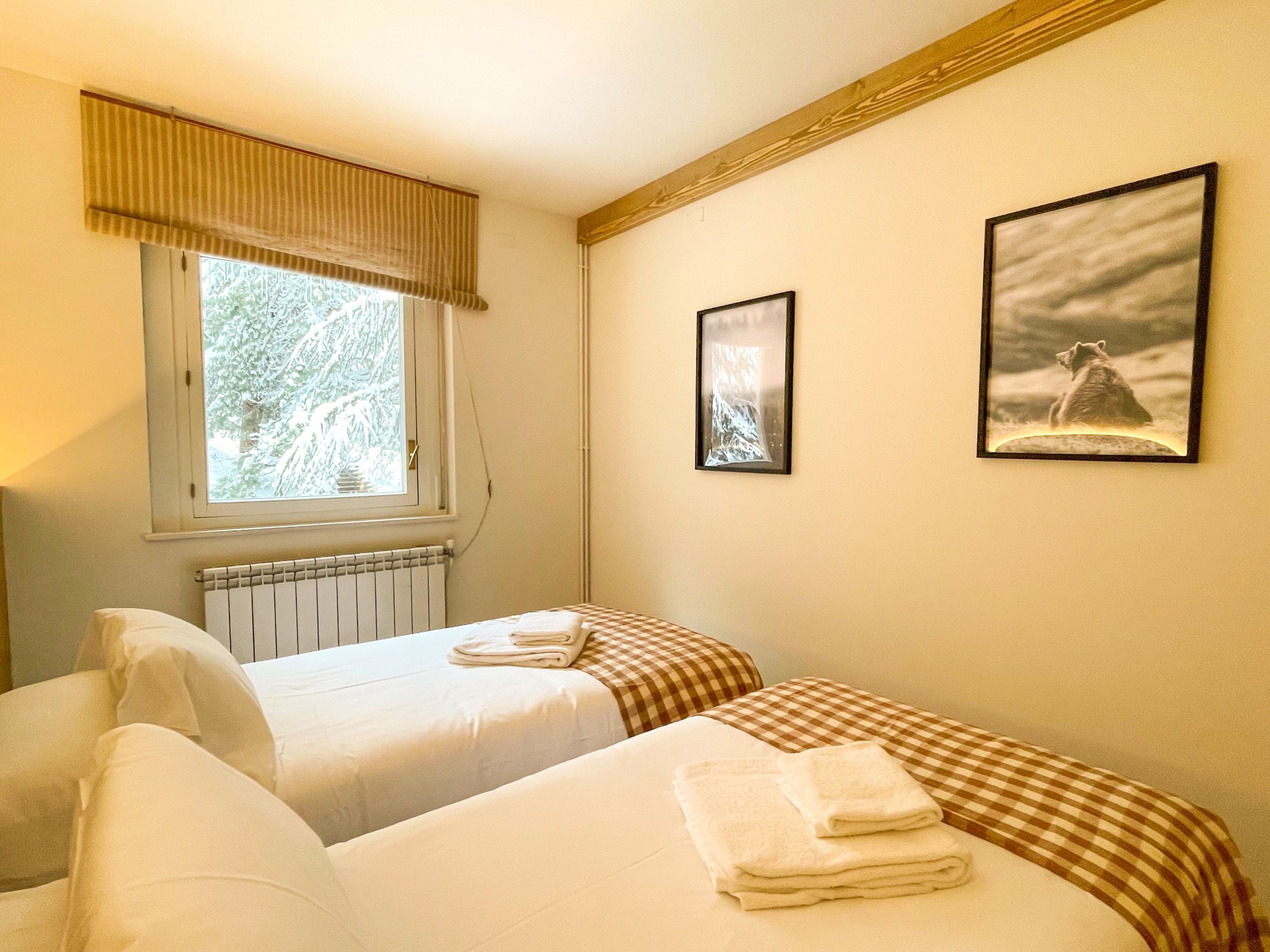 A hotel room with two beds, a window with a view of trees, a radiator, and two framed pictures on the wall.