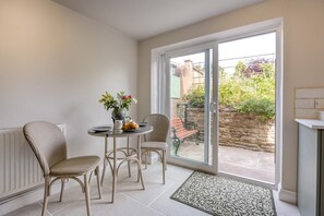 Ein sonnendurchflutetes Zimmer mit einem für Tee gedeckten Tisch, zwei Korbstühlen und Blick auf einen Garten durch eine Glastür, außerdem eine Steinmauer im Freien.