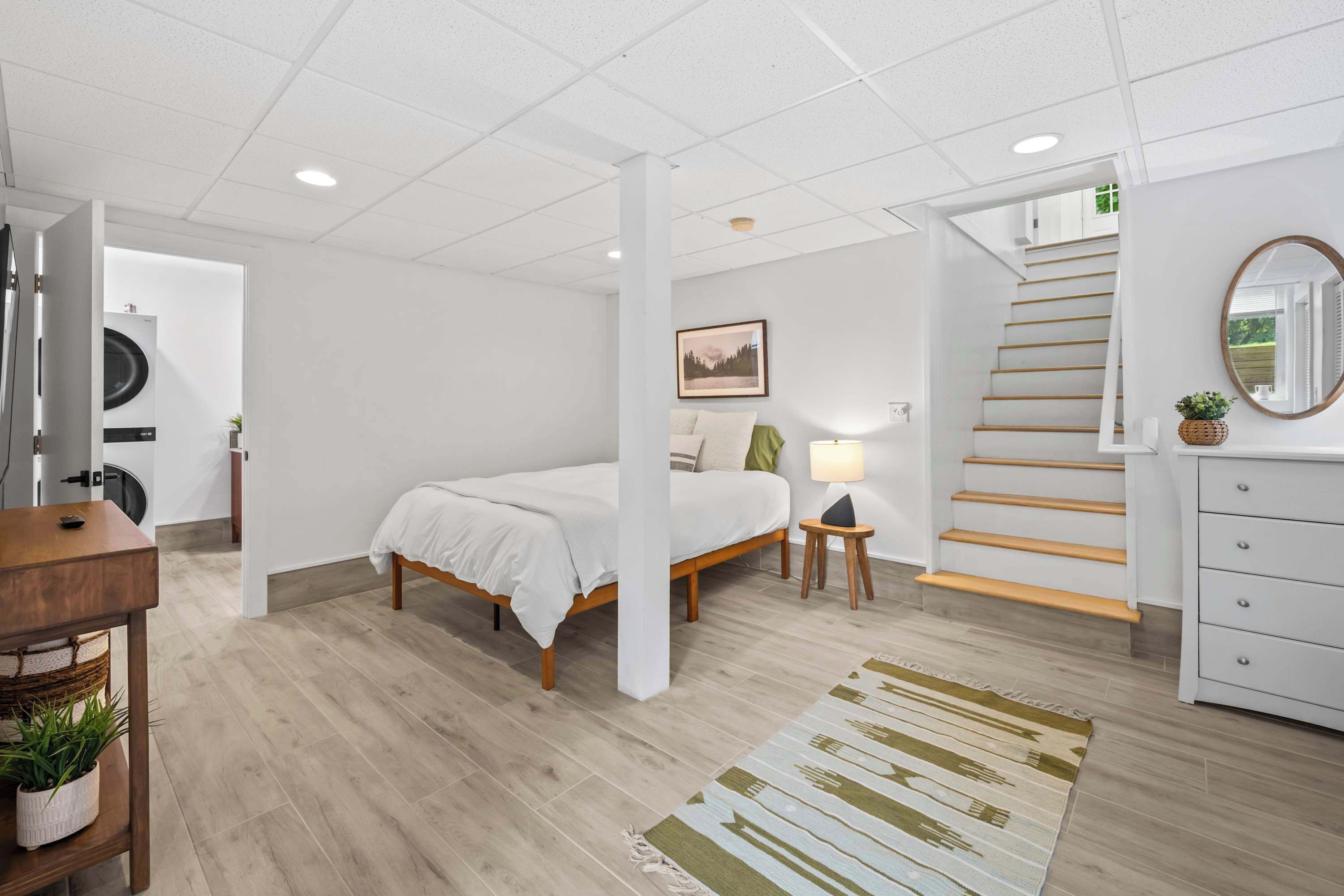 Ein Schlafzimmer mit einem Bett, Nachttischen, einer Lampe, einem Schminktisch, einem Spiegel, einer Treppe und einer Waschmaschine im Hintergrund.