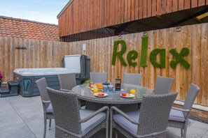 An outdoor patio with a hot tub, wicker chairs, and a table set for breakfast. A wooden fence with a 'Relax' sign is in the background. - 4 Bed in Skirlaugh (oc-w43303) (Skirlaugh)