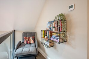 A corner of a room with a grey cushioned chair, a small bookshelf with books and a potted plant, and a thermostat on the wall. - 4 Bed in Skirlaugh (oc-w43303) (Skirlaugh)