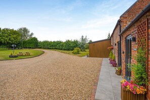 A gravel driveway leading to a building with a brick facade, a wooden bench, and flower planters. - 4 Bed in Skirlaugh (oc-w43303) (Skirlaugh)