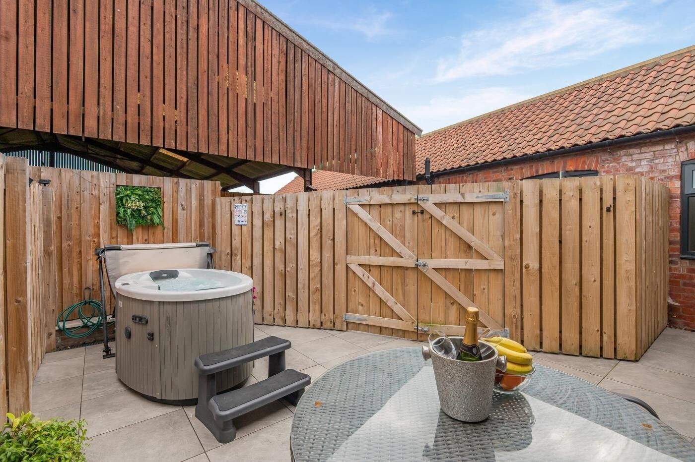 A backyard with a hot tub, a wooden fence, a brick wall, and a table with a bottle of champagne.