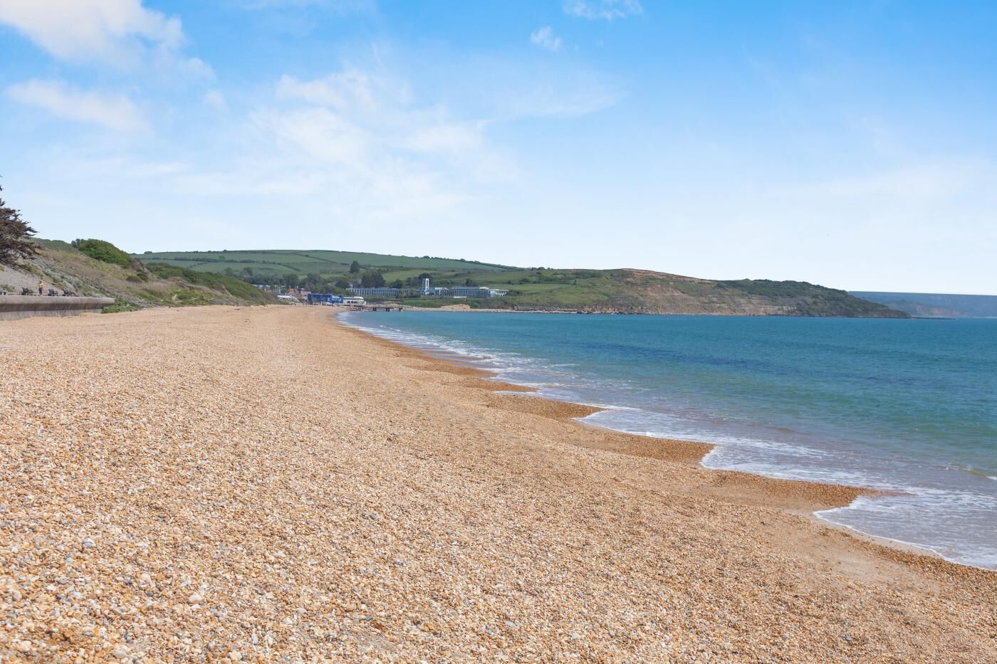 A pebble beach with a calm sea, distant hills, and a few buildings.