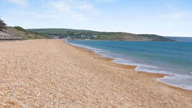 A pebble beach with a calm sea, distant hills, and a few buildings. - 2 Bed in Weymouth (oc-s42788) (Weymouth)