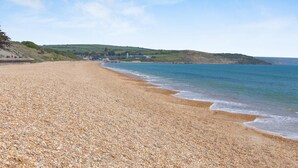 A pebble beach with a calm sea, distant hills, and a few buildings. - 2 Bed in Weymouth (oc-s42788) (Weymouth)