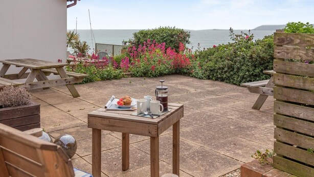 A patio with a wooden table set for breakfast, a view of the sea, and a wooden fence. - 2 Bed in Weymouth (oc-s42788) (Weymouth)