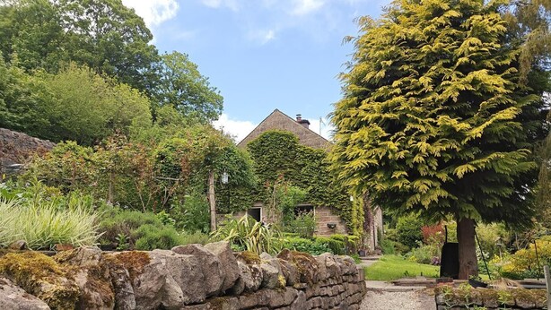 A stone wall leading to a rustic stone building surrounded by lush greenery and trees. - 3 Bed in Whatstandwell (oc-h42743) (Whatstandwell)