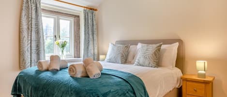 A neatly made bed with a teal bedspread, two pillows, and rolled towels on a wooden nightstand with a lamp.