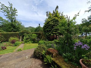 A garden with a stone pathway, various plants, and a white vehicle parked in the background. - 3 Bed in Whatstandwell (oc-h42743) (Whatstandwell)