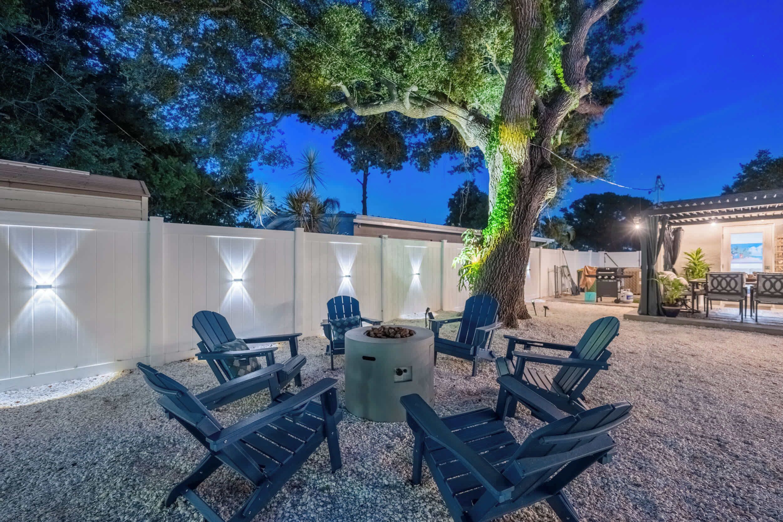 An outdoor seating area with Adirondack chairs and a fire pit, surrounded by a white fence and trees.