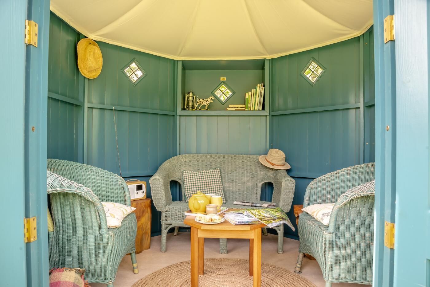 A cozy room with a checkered armchair, wicker chairs, a wooden table with a teapot, and a radio.