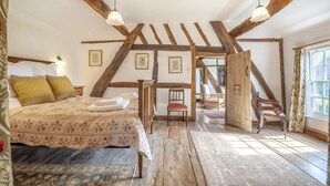 Ein Schlafzimmer mit Holzbalkendecke, einem Bett mit floralem Bettzeug, einem Sessel und einem Fenster mit Vorhängen.