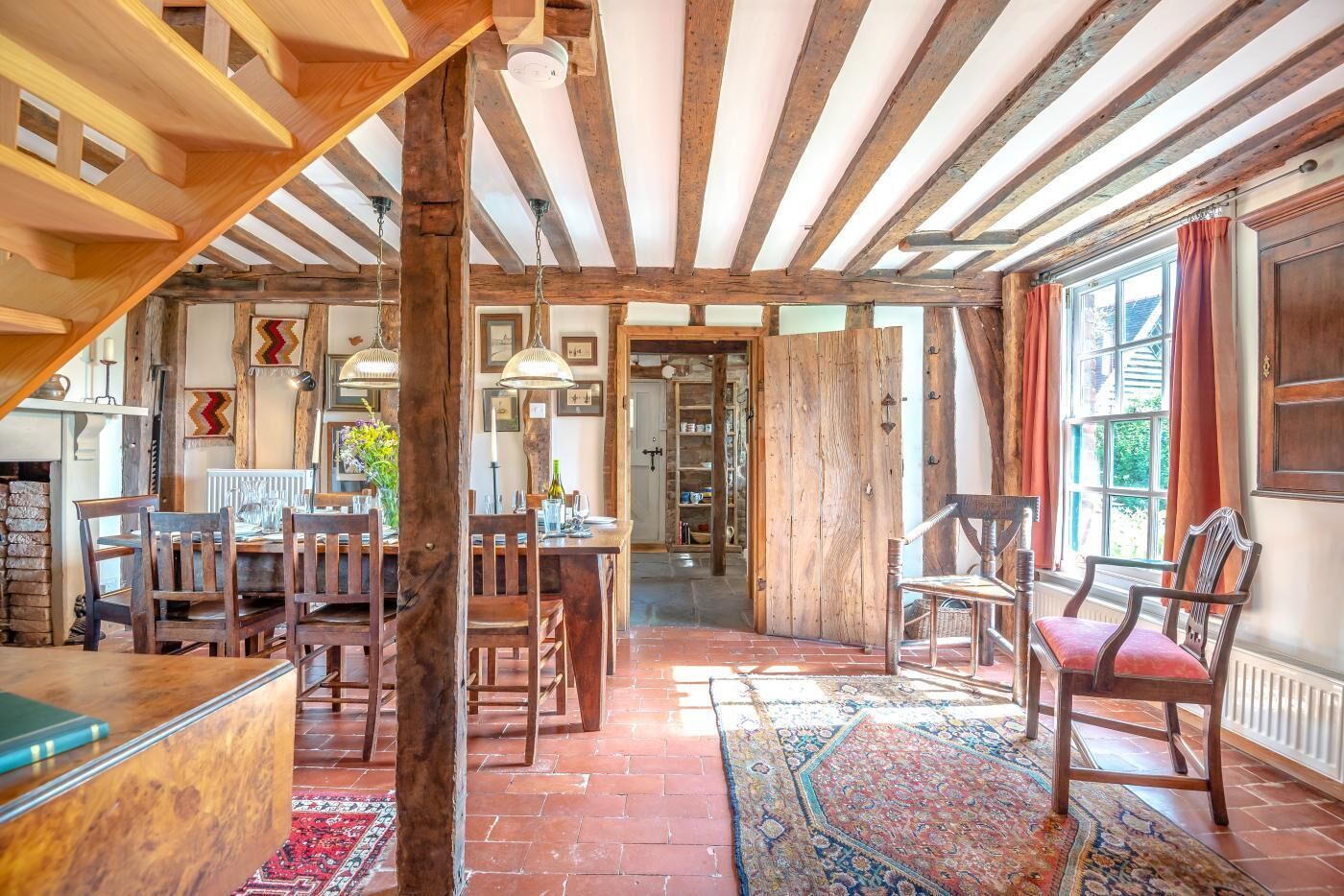 Un interior rústico con vigas de madera a la vista, una zona de comedor con sillas de madera, una alfombra estampada y una vista a través de un vano.