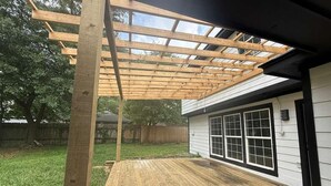 A wooden deck with a pergola structure, a covered patio area, and a house with a large window. - Spacious 4BR Home · Near IAH · Self Check-In (Spring)